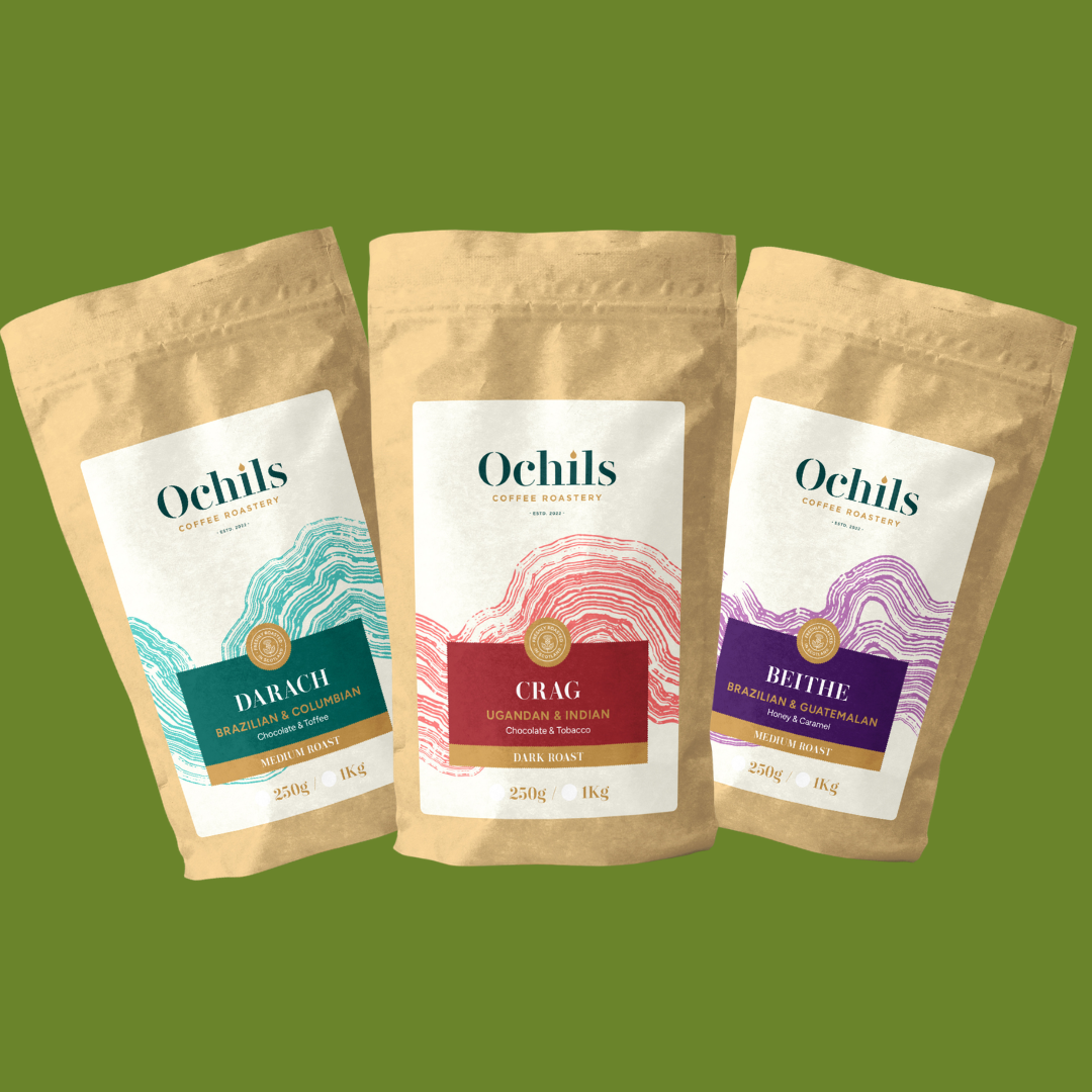 Three stand-up pouches of Ochils coffee beans arranged side by side on a solid green background. The pouches are kraft paper with white labels featuring different accent colors: teal, red, and purple, with the flavors Darach, Crag, and Beithe.